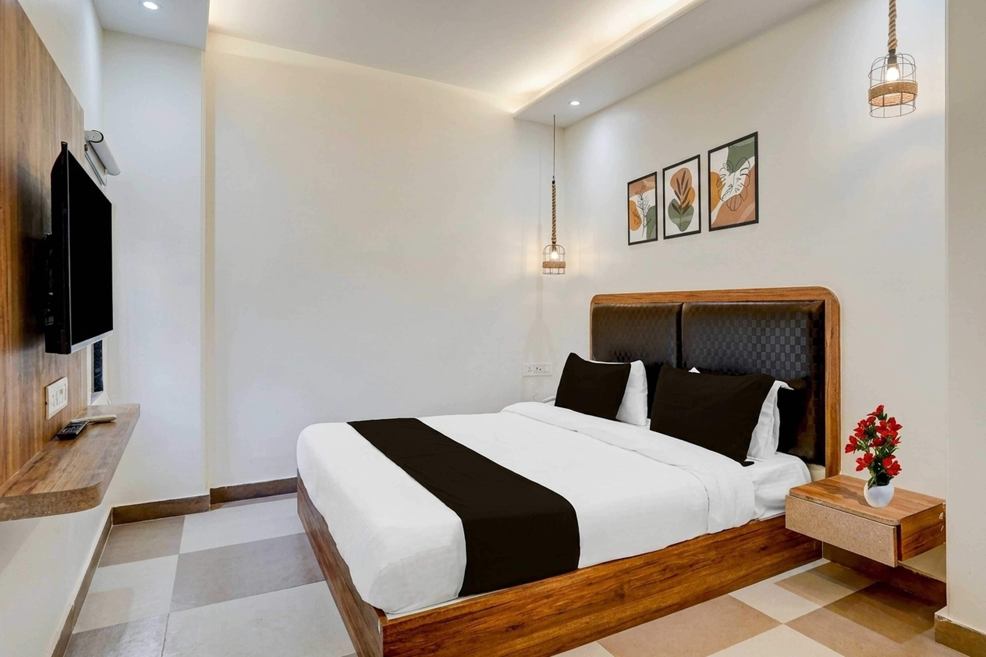 Una habitación de hotel moderna con una cama grande, cabecera de madera, una mesita de noche con un jarrón de flores y un televisor de pantalla plana montado en la pared.