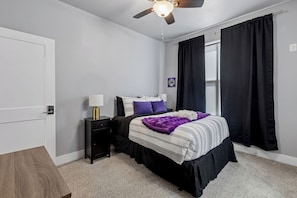 A bedroom with a bed, a nightstand, a lamp, a ceiling fan, and black curtains.