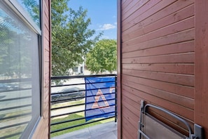 A view from a window to a balcony with a metal railing, a blue and red gate, and a tree-lined street in the background.