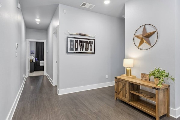 A hallway with a sign that reads 'HOWDY' on the wall, a star decoration, and a wooden console table with a lamp and a plant.