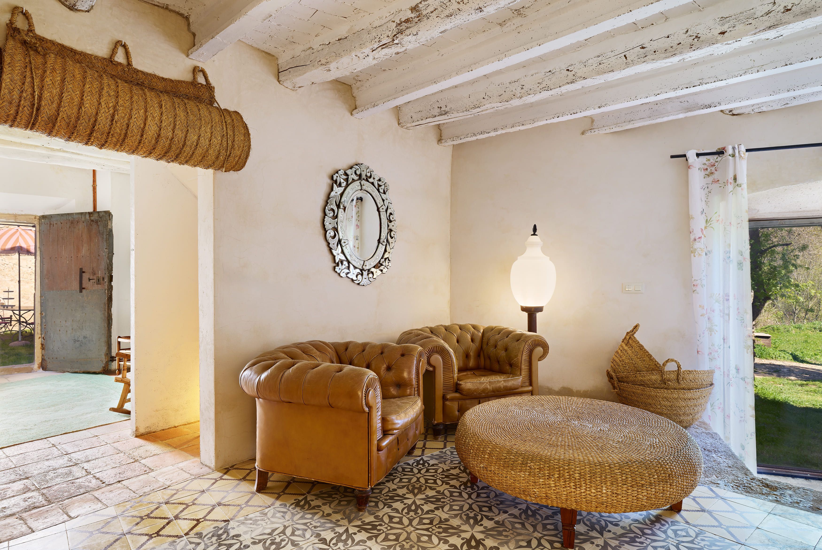 A room with a patterned floor, two leather sofas, a woven pouf, a wall-mounted mirror, and a hanging basket.