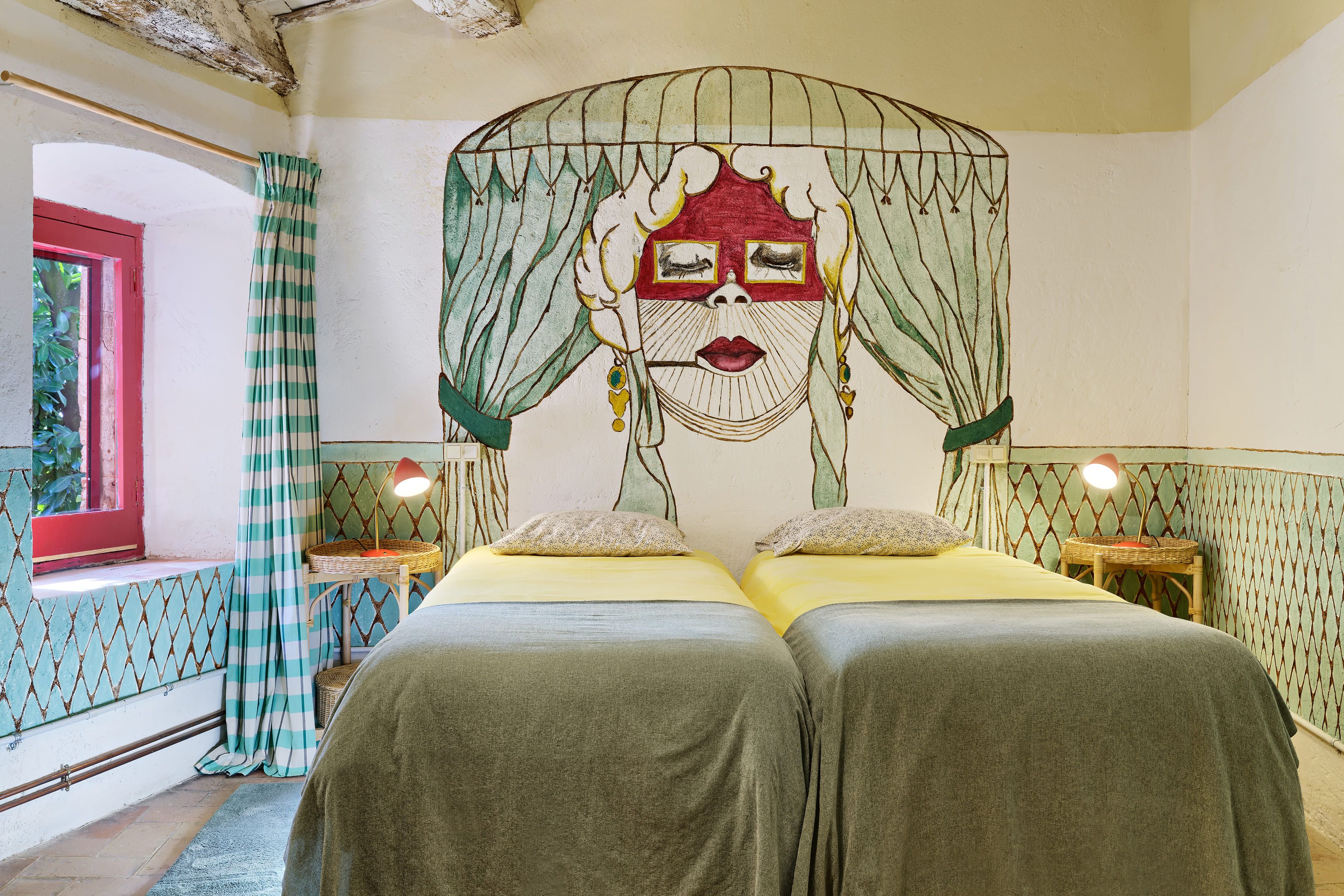 A room with two beds, a window with curtains, a wall with a painted design, and a small table with a lamp.