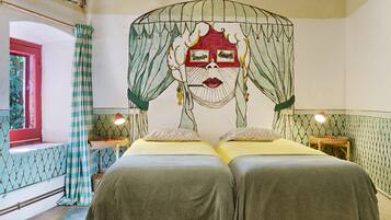 A room with two beds, a window with curtains, a wall with a painted design, and a small table with a lamp.