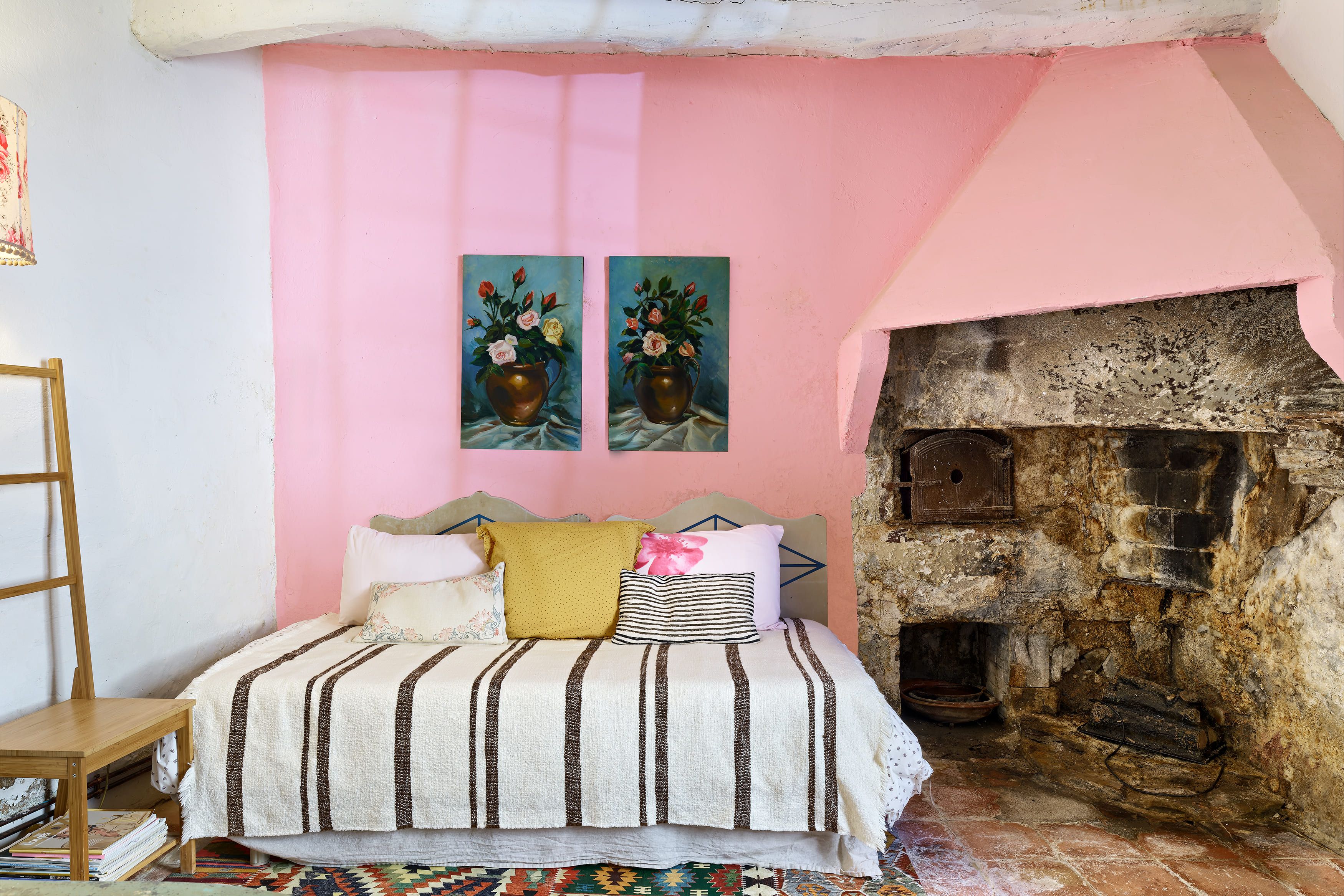 Une chambre à coucher avec un lit, des oreillers, une échelle en bois, une cheminée et deux tableaux accrochés au mur.