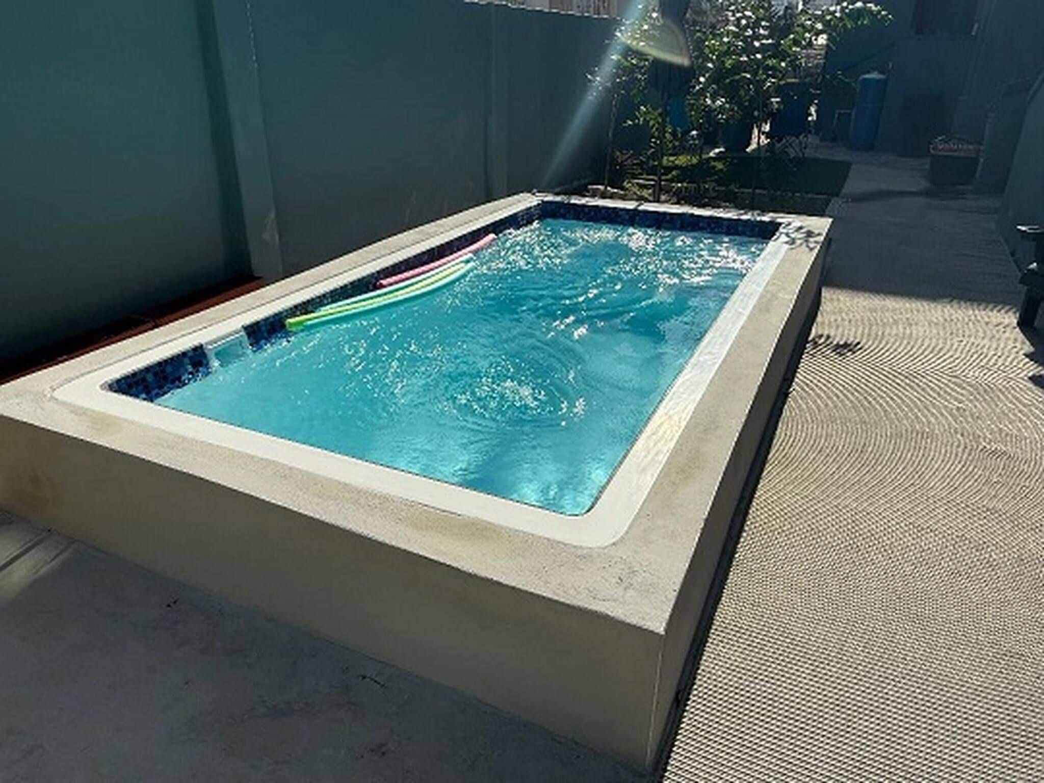 Un bain à remous extérieur rectangulaire, avec une eau d’un bleu limpide, entouré d’une terrasse en béton et d’un tapis texturé.