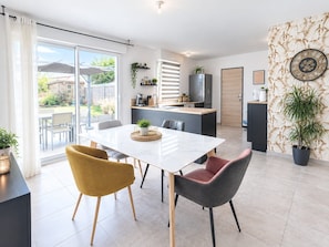 A modern kitchen with a central island, a dining area with a white table and two chairs, and a view of a garden through a sliding door. - La Parenthèse de Chaumes  Proche de la mer  T3 (Chaumes-en-Retz)