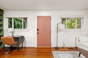 A room with a wooden floor, a desk with a chair, a white sofa, a pink door, and two windows with greenery outside. - Cozy Broad Ripple Bungalow | Walk to Everything (Indianapolis)