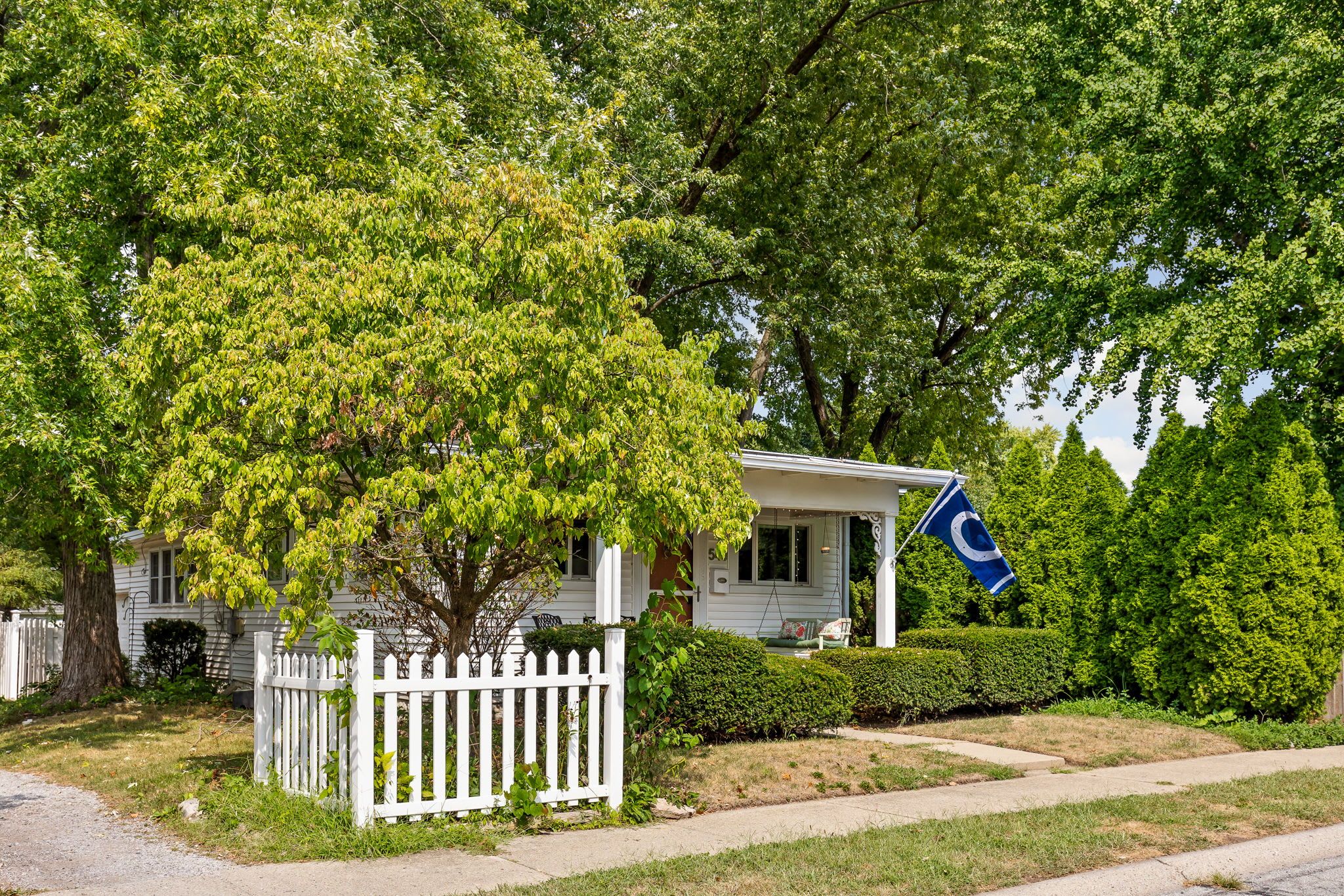 A residential house with a white picket fence, a tree in front, and a flag on the porch.
