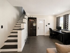 Ein Wohnesszimmer mit Treppe, Schreibtisch inklusive Computer und Bücherregal.