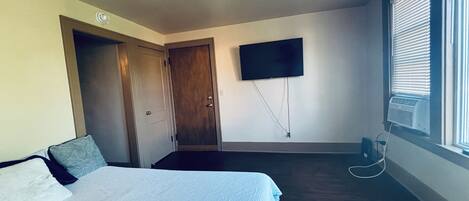 A room with a bed, a flat-screen TV mounted on the wall, a window with blinds, and a door.