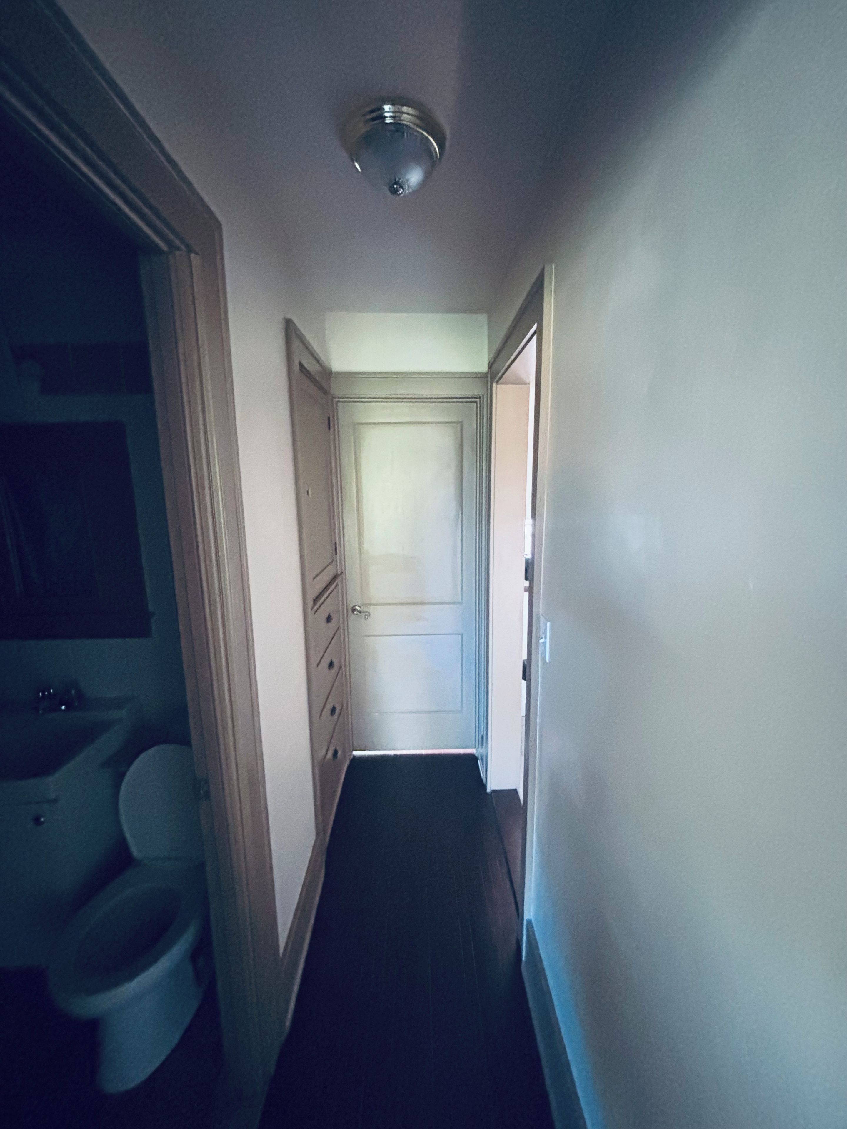 Ein schmaler Flur mit einer Tür am Ende, einer Deckenleuchte und einem Badezimmer mit Toilette und Waschbecken auf der linken Seite.