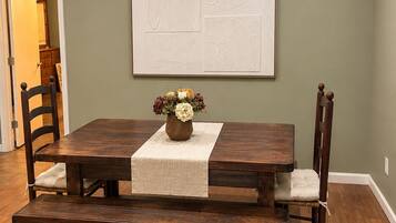 A dining area with a wooden table, cushioned chairs, a vase with flowers, and a wall-mounted artwork.