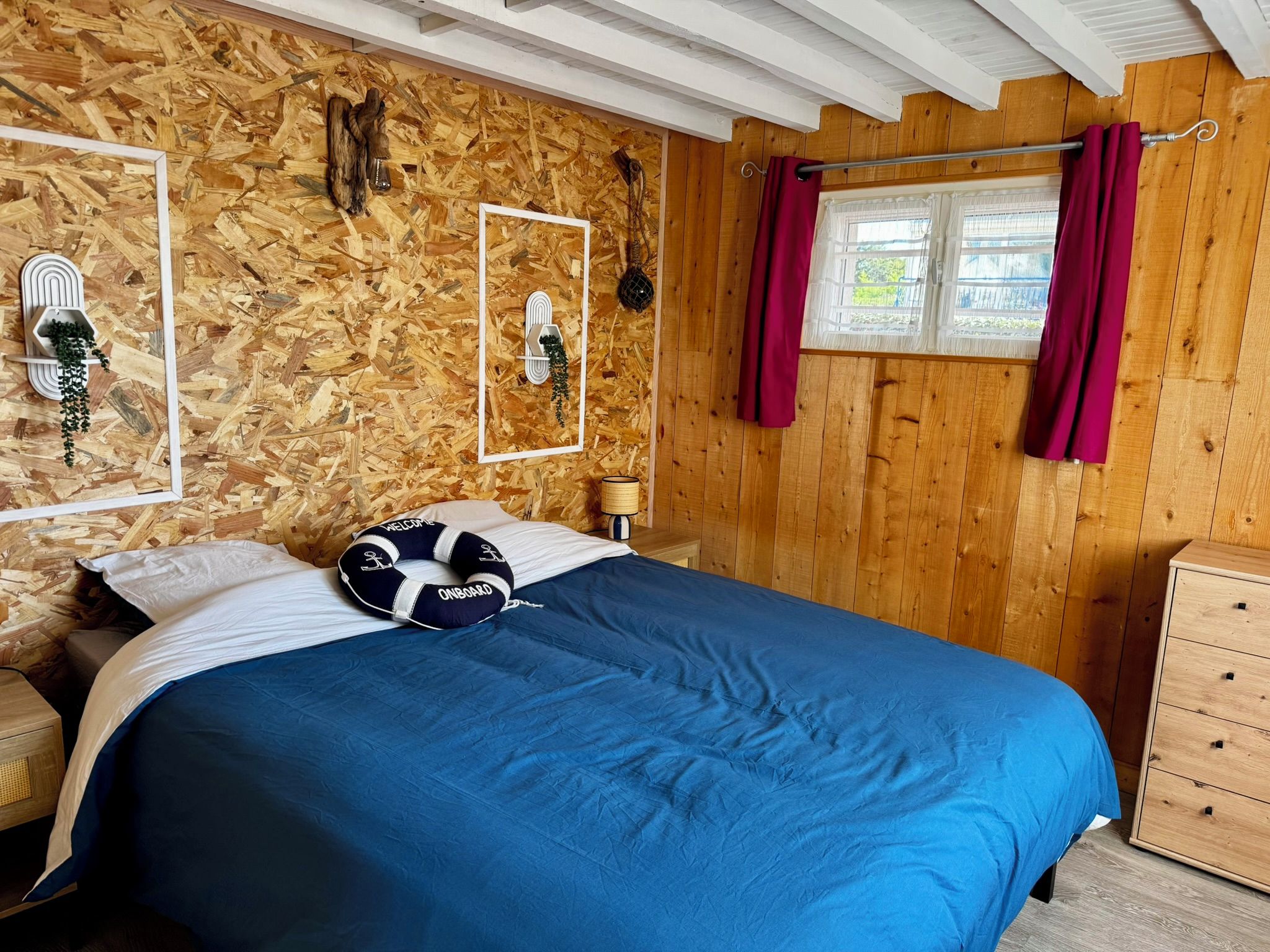 A bedroom with a wooden wall, a bed with a blue bedspread, a life preserver pillow, a window with curtains, and a wooden dresser.