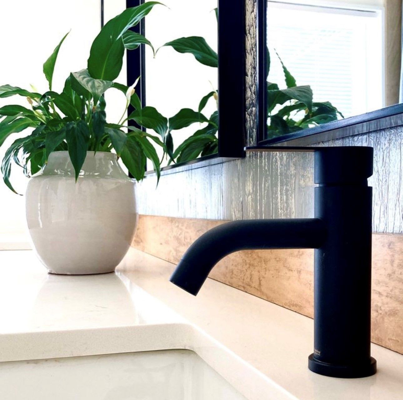 Een moderne badkamer met een zwarte kraan, een witte vaas met groene planten, en een raam met luxaflex.