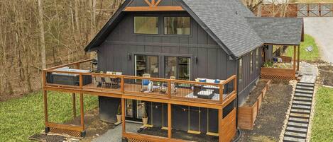A modern two-story house with a balcony and a deck, surrounded by a forest.