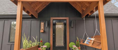 A wooden porch with a swing chair, potted plants, and a door with a number sign.
