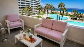 Ein Balkon mit einer Couch und Sesseln, einem kleinen Tisch mit Snacks und Getränken sowie Blick auf einen Pool und Strand.