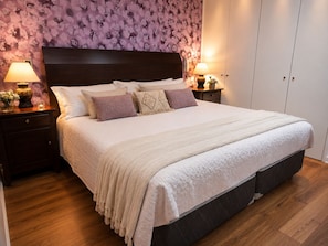 Ein ordentlich bezogenes Bett mit weißer Daunendecke und mehreren Kissen, ein hölzernes Kopfteil, ein Nachttisch mit Lampe und eine Wand mit Blumenmuster.