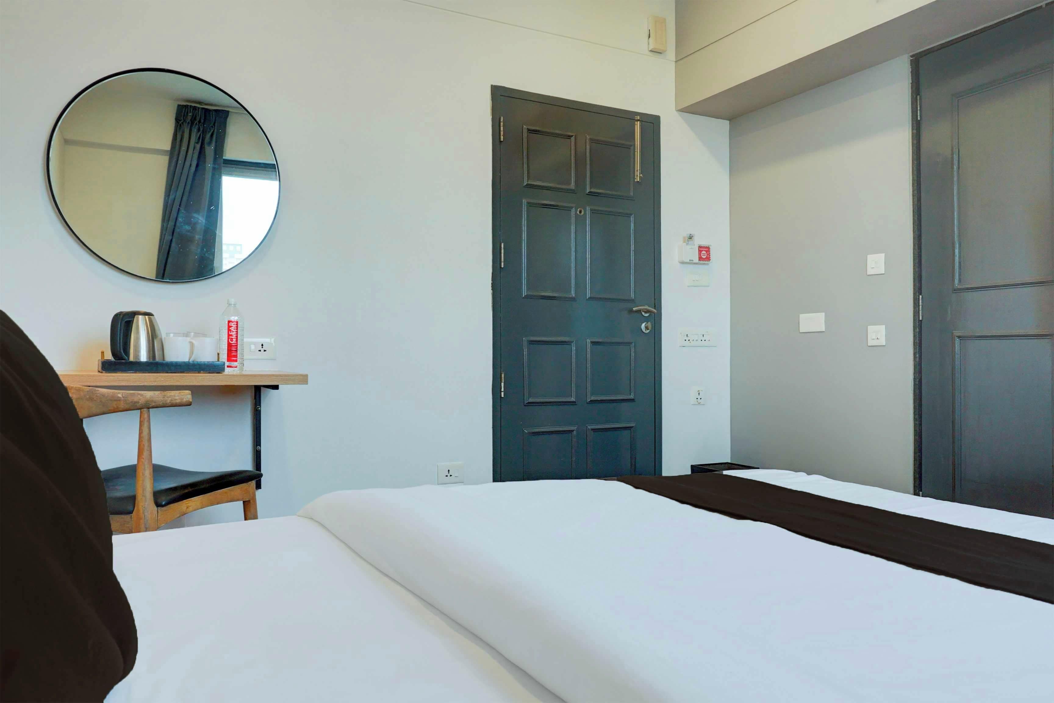A hotel room with a bed, a desk with a kettle, a mirror, and a door.