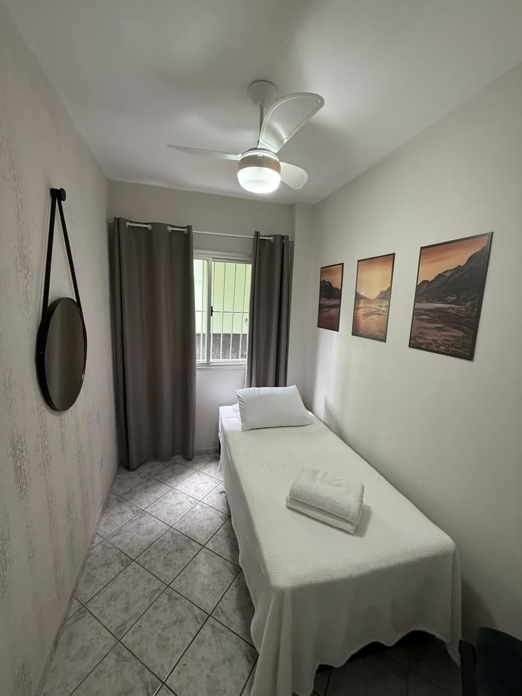 A small, single-bed room with a ceiling fan, a window with curtains, and three landscape paintings on the wall.