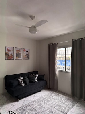 Una sala de estar con un sofá negro, un ventilador de techo, un ventanal con cortinas y tres obras de arte colgadas en la pared.