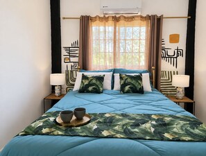 A bed with a blue quilt, two bedside lamps, a window with curtains, and a wall with abstract art. - Stylish Sanctuary: Mountain & Sea Views at Nya’s Place from Your Private Balcony (Tower Isle)