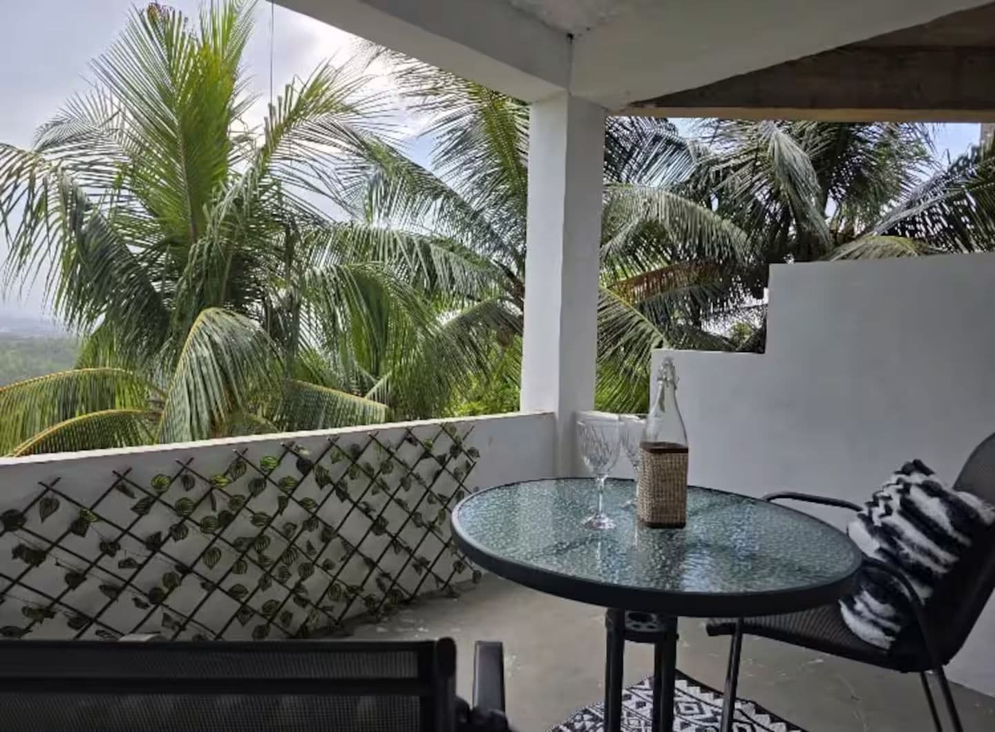 A glass-top table with a bottle and glass, a black and white patterned chair, and a view of palm trees.