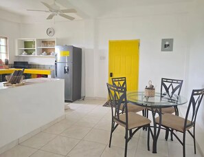 A modern kitchen with a refrigerator, a dining table with chairs, and a yellow door. - Stylish Sanctuary: Mountain & Sea Views at Nya’s Place from Your Private Balcony (Tower Isle)