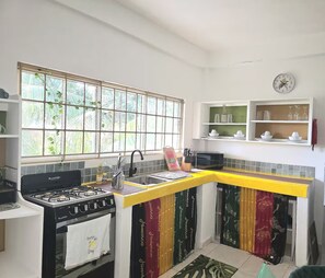 Une cuisine équipée d’une cuisinière à gaz, d’un évier et d’une fenêtre agrémentée de plantes. On y trouve des étagères remplies d’ustensiles de cuisine et une horloge au mur.