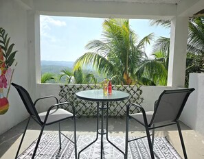 Un balcon avec une table et des chaises, donnant sur un paysage tropical avec des palmiers et une vue sur l’océan.