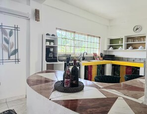 A modern kitchen with a curved island, a coffee machine, and a window with a view of greenery. - Stylish Sanctuary: Mountain & Sea Views at Nya’s Place from Your Private Balcony (Tower Isle)