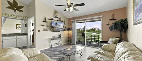A living room with a sofa, a coffee table, and a ceiling fan. There is a television on the wall and a balcony with a view.