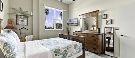 Une chambre à coucher comprenant un lit, une commode, un miroir, un ventilateur de plafond et une fenêtre donnant sur des palmiers.