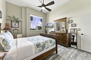 A bedroom with a bed, a dresser, a mirror, a ceiling fan, and a window with a view of palm trees. - NEW Golf Course View 2BR Condo | Isla Del Sol | St Pete (Isla Del Sol)