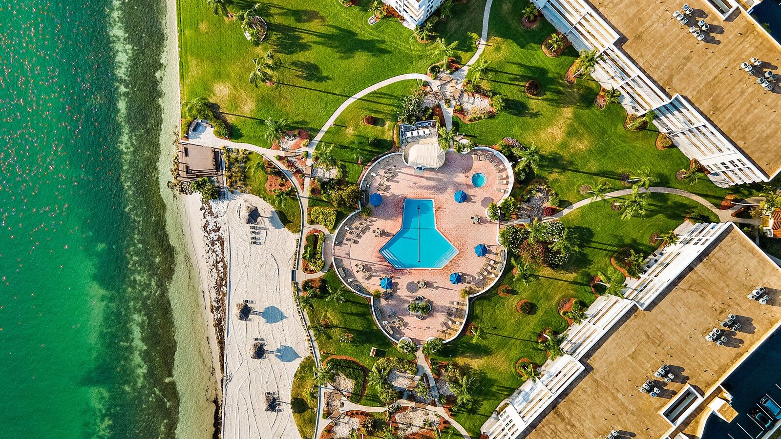Vista aérea de un complejo turístico con una piscina central, varias edificaciones y una zona de playa.
