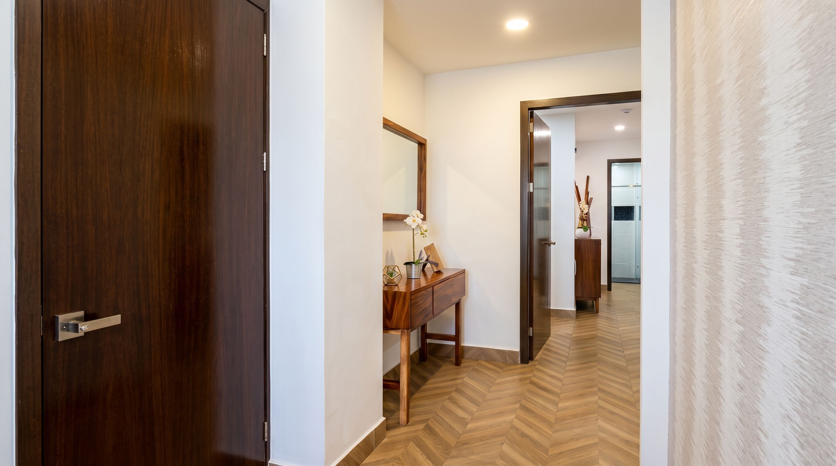 A hallway with a wooden door, a mirror, a wooden console table with a vase, and a herringbone-patterned floor.