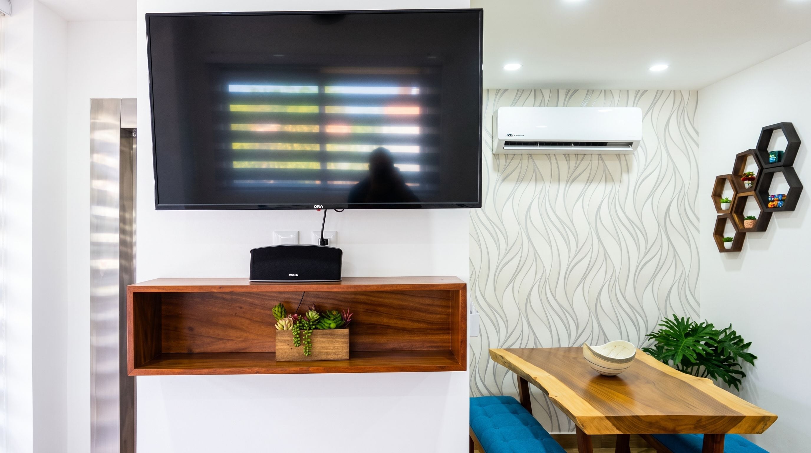 Un salon moderne avec un téléviseur à écran plat fixé au mur, une console en bois agrémentée d’objets décoratifs, une table à manger avec un saladier, et un appareil de climatisation.