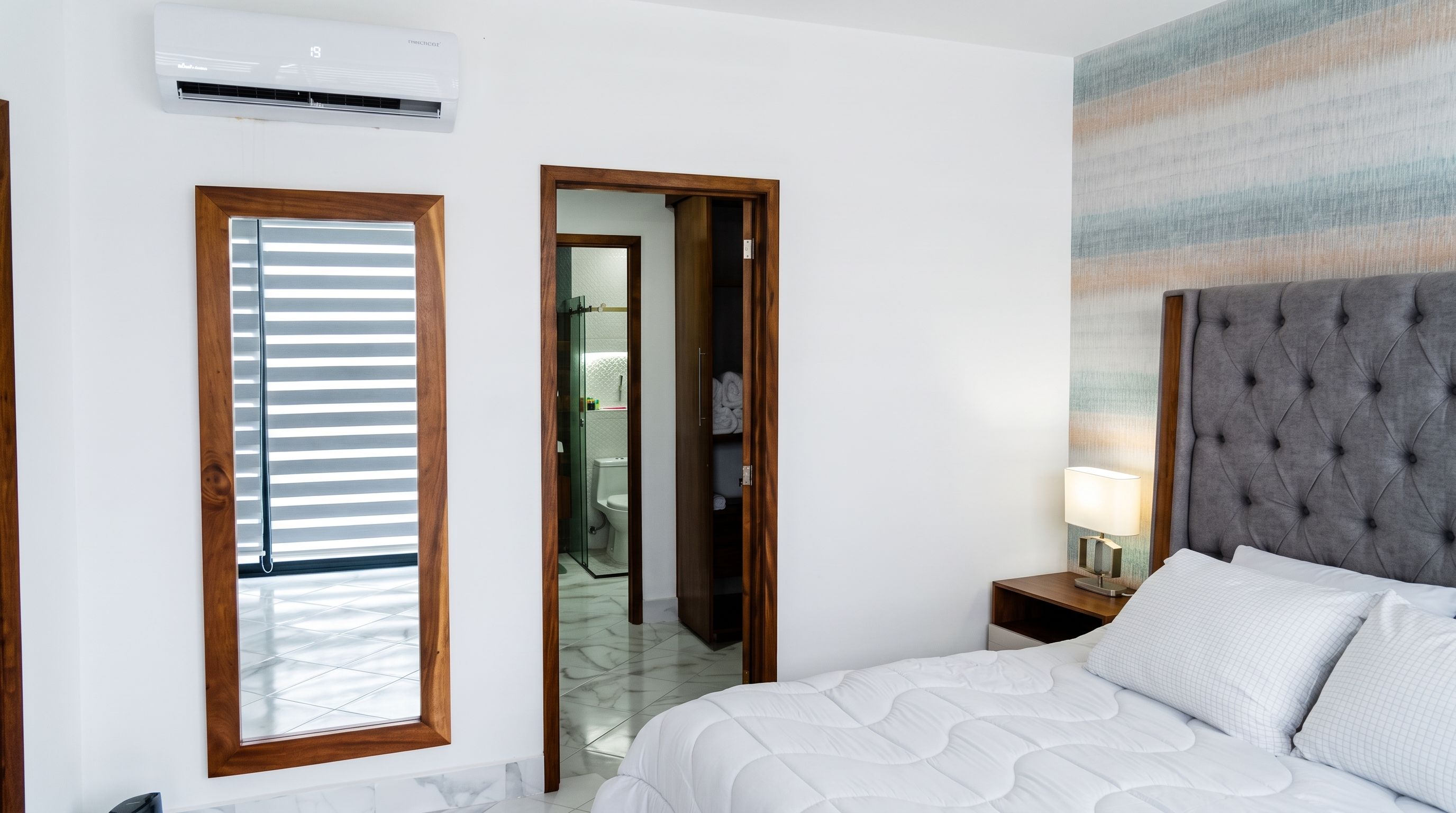 Une chambre d’hôtel comprenant un lit, une table de chevet, une lampe, un appareil de climatisation, une fenêtre avec des stores et une salle de bain visible à travers une porte ouverte.