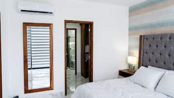 Une chambre d’hôtel comprenant un lit, une table de chevet, une lampe, un appareil de climatisation, une fenêtre avec des stores et une salle de bain visible à travers une porte ouverte.