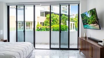 A modern bedroom with a large bed, a TV mounted on the wall, and a sliding glass door leading to a balcony with a view of greenery and buildings.