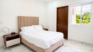 A bedroom with a bed, nightstands, a wooden door, and a window with a view of greenery.