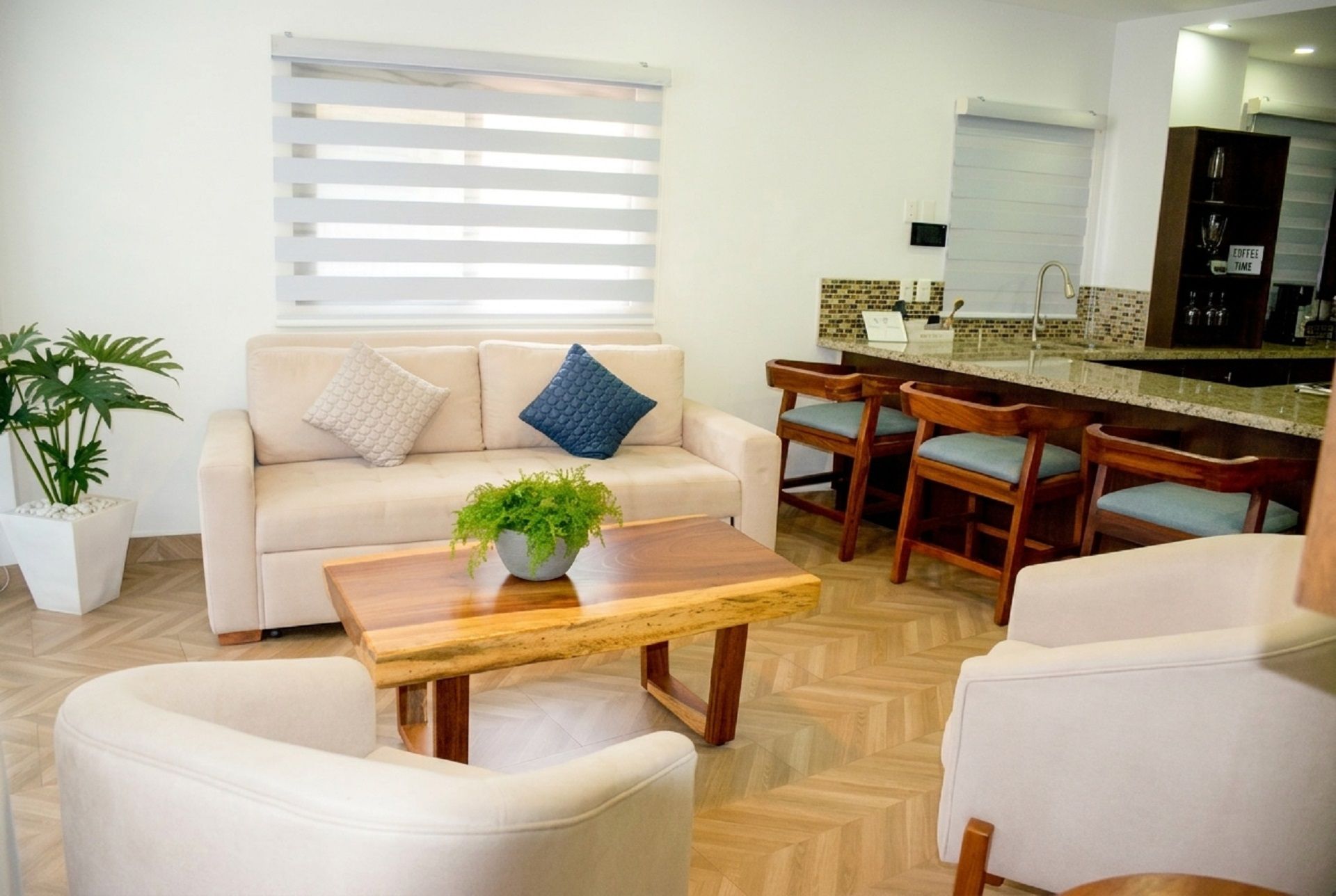 Una sala de estar con un sofá, una mesa de centro con una planta, y un comedor con sillas de madera, con una cocina al fondo.