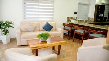 Una sala de estar con un sofá, una mesa de centro con una planta, y un comedor con sillas de madera, con una cocina al fondo.