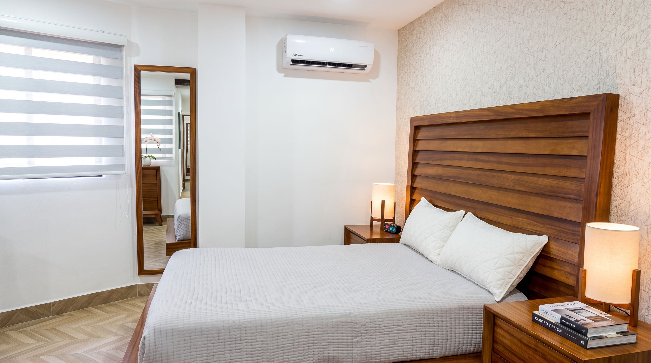 Habitación de hotel con cama, lámparas de noche, mesita de noche, aire acondicionado instalado en la pared y ventana con persianas.