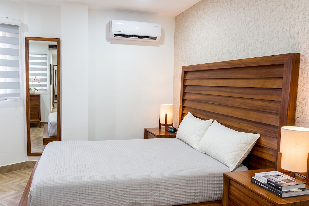 Habitación de hotel con cama, lámparas de noche, mesita de noche, aire acondicionado instalado en la pared y ventana con persianas.