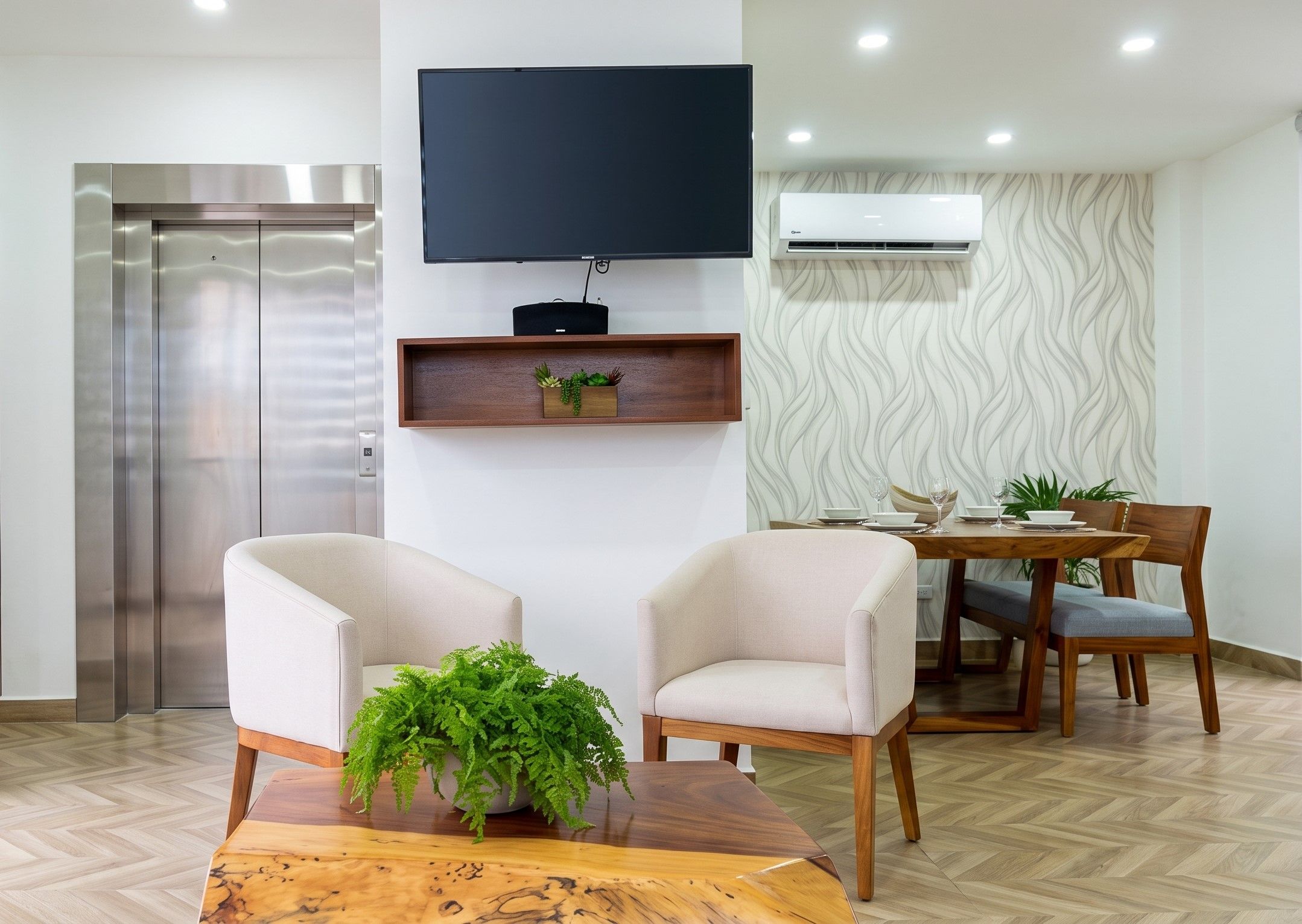 A modern living room with a flat-screen TV mounted on the wall, a wooden coffee table with a plant, two armchairs, and a dining area with a wooden table and chairs.