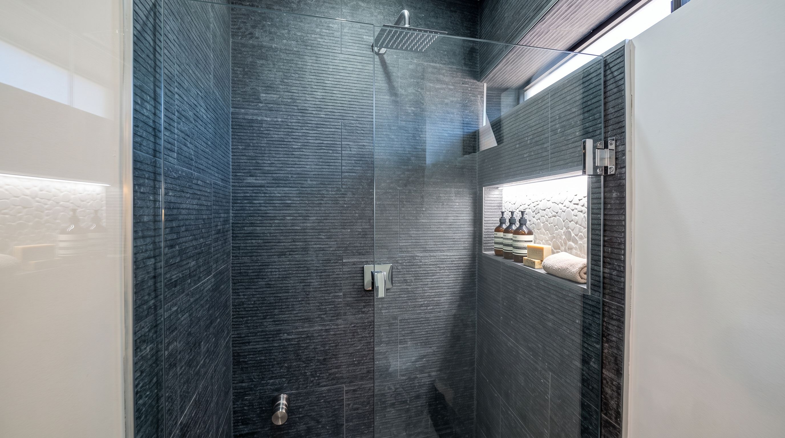 Un baño moderno con paredes de azulejos oscuros, cabina de ducha con mampara de vidrio y repisa con artículos de aseo.