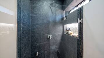 Un baño moderno con paredes de azulejos oscuros, cabina de ducha con mampara de vidrio y repisa con artículos de aseo.
