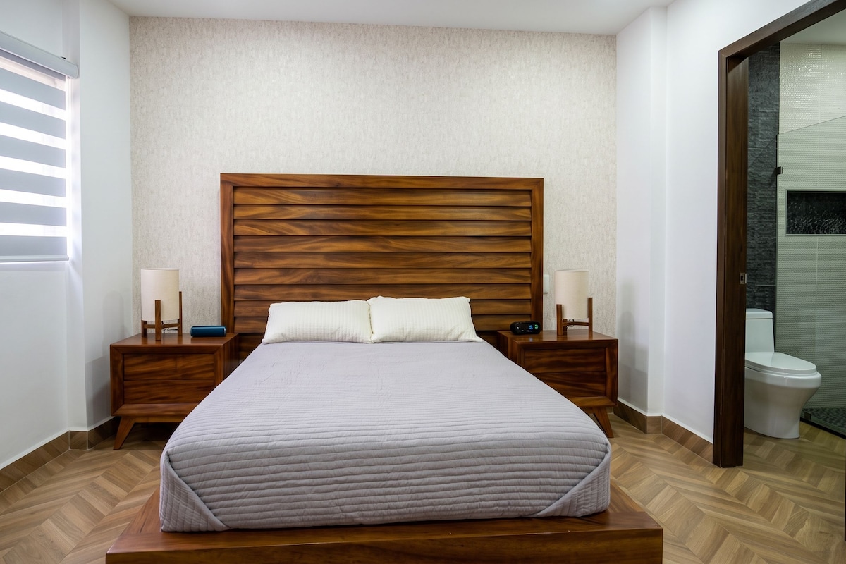 Una cama bien tendida con cabecera de madera, dos mesitas de noche con lámparas, y vista a un baño por una puerta abierta.