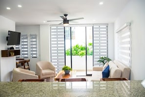A modern living room with a flat-screen TV, a ceiling fan, and a sliding glass door leading to a balcony with greenery.